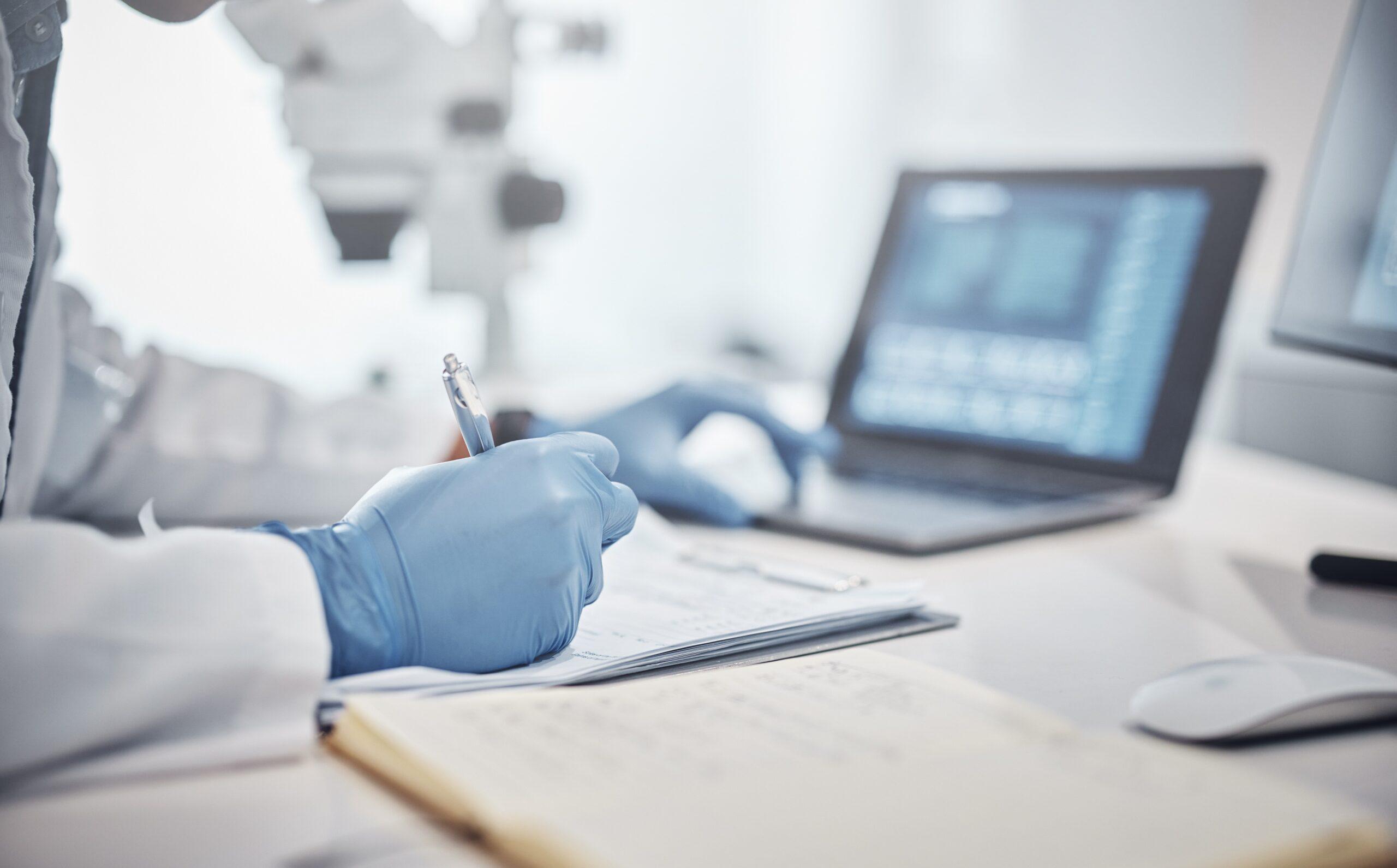 Stock of a medical professional taking notes at a computer and on a paper.