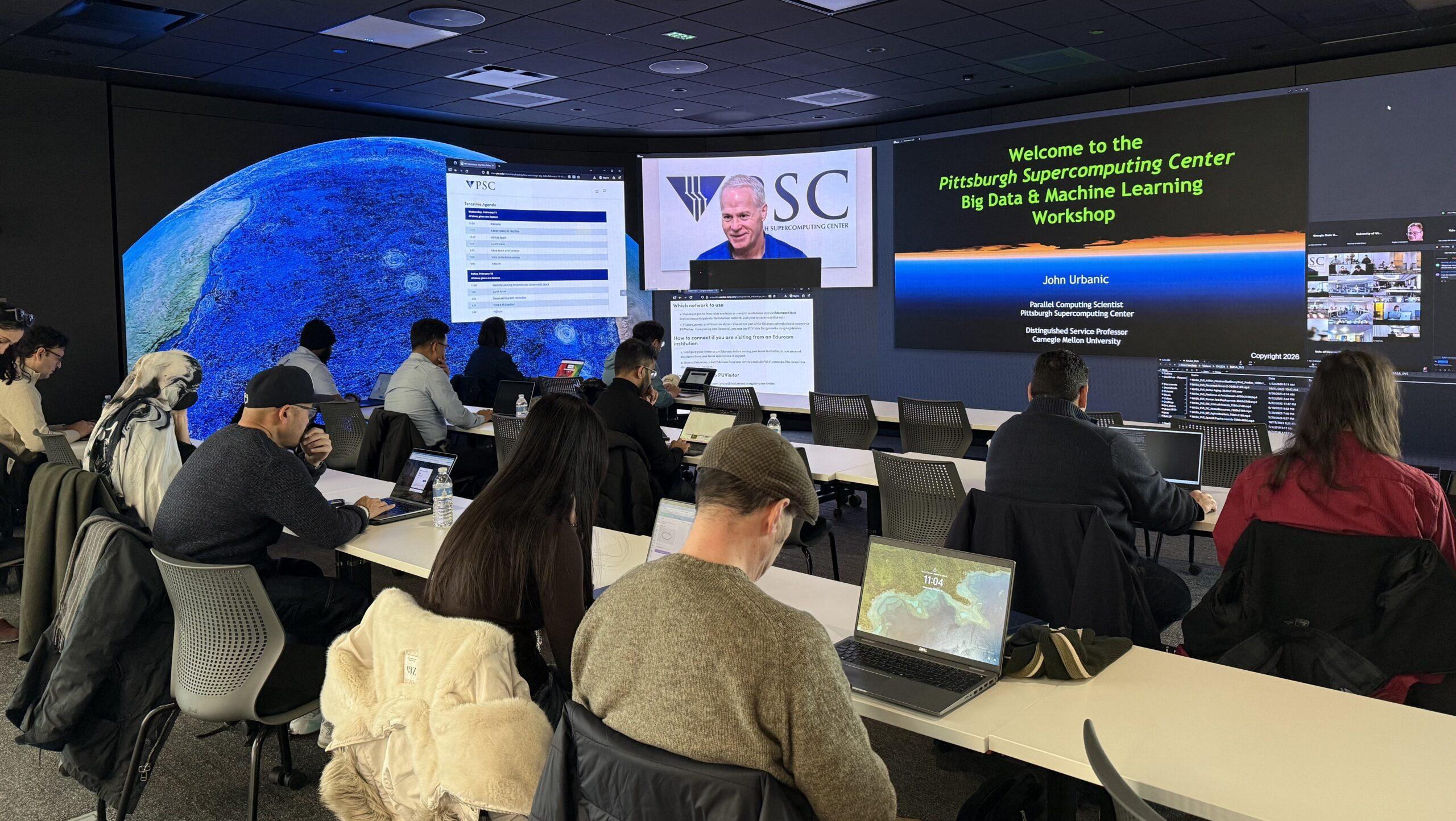 A man speaks on a screen to a classroom of students at computers