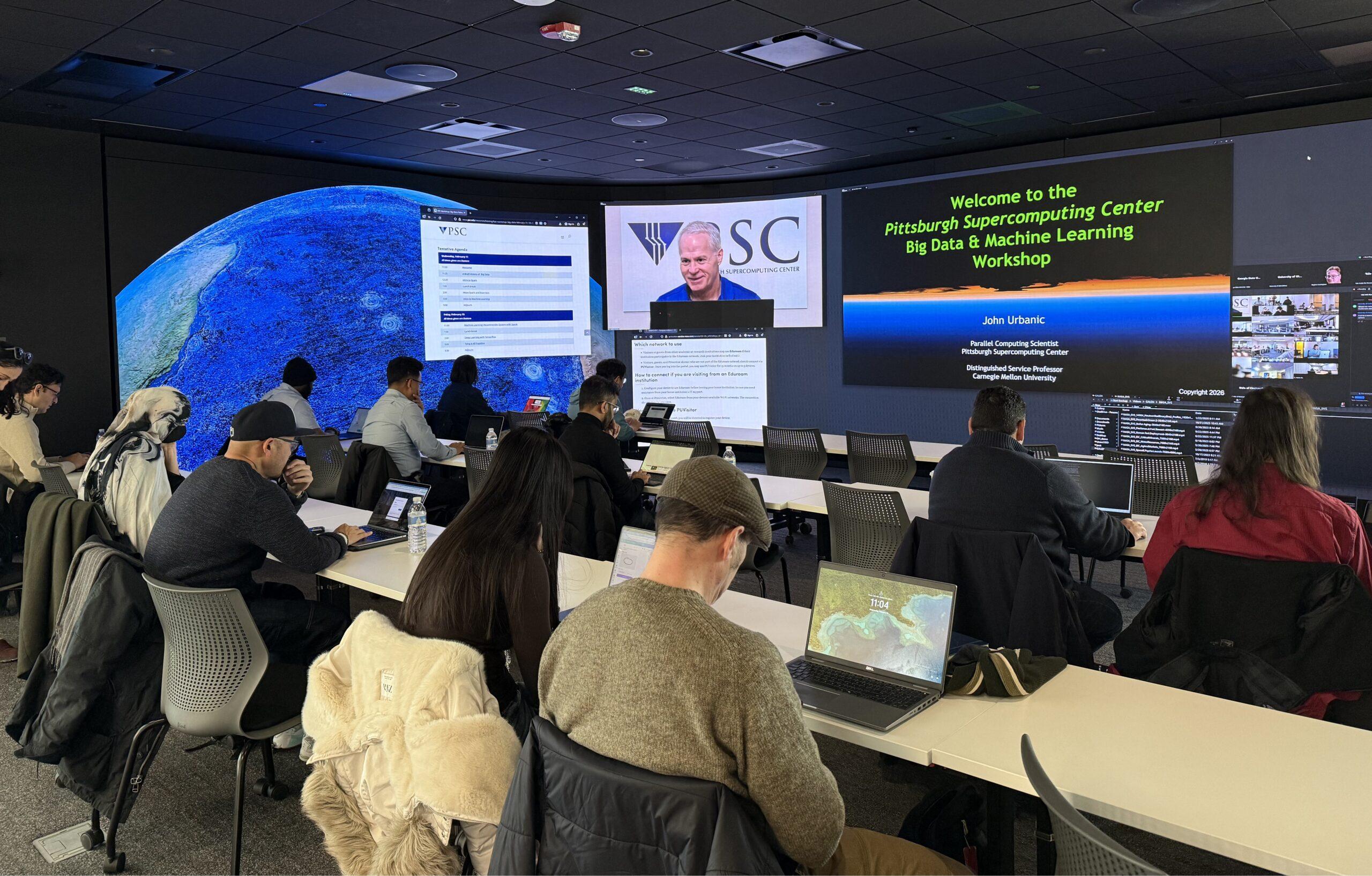 A man speaks on a screen to a classroom of students at computers