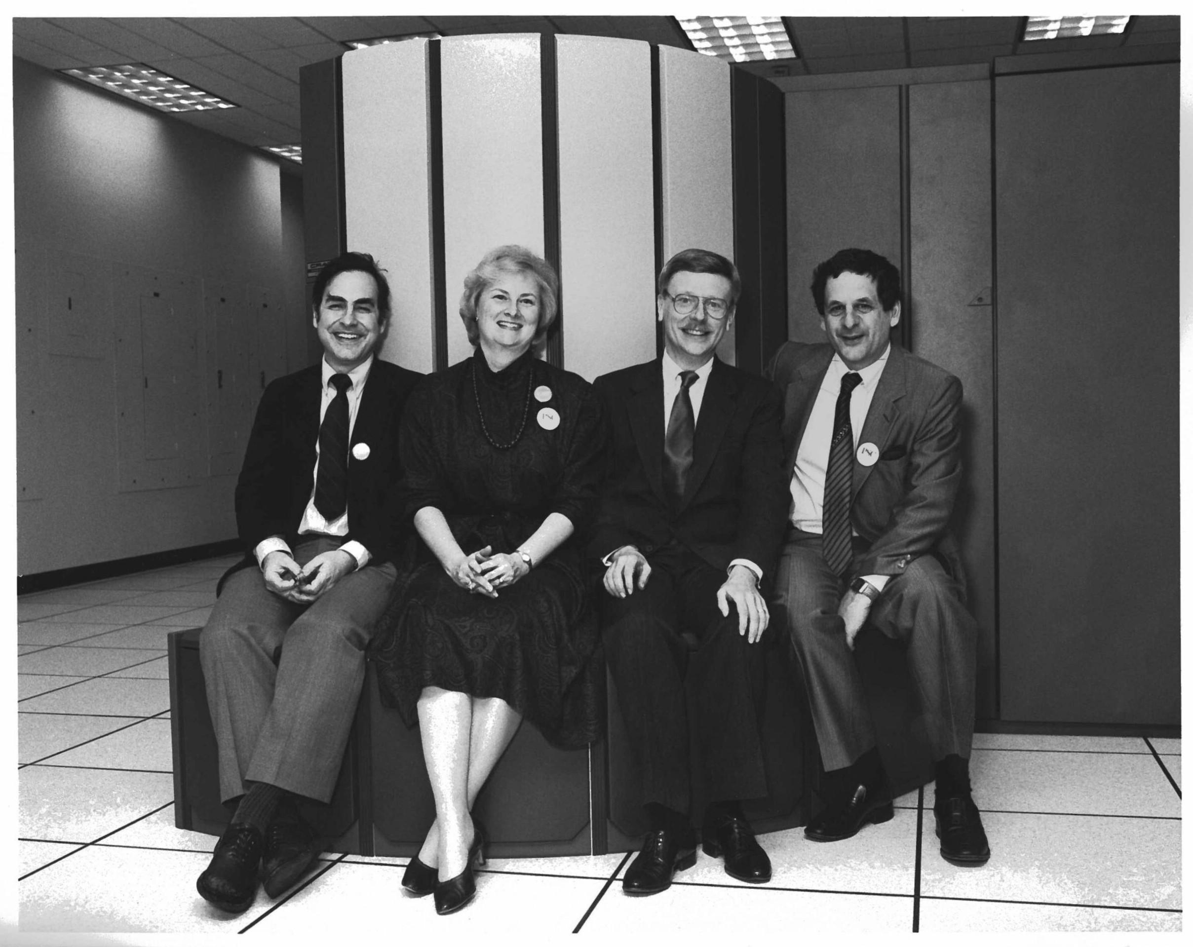 Four people sit on the bench of a 1980s supercomputer.