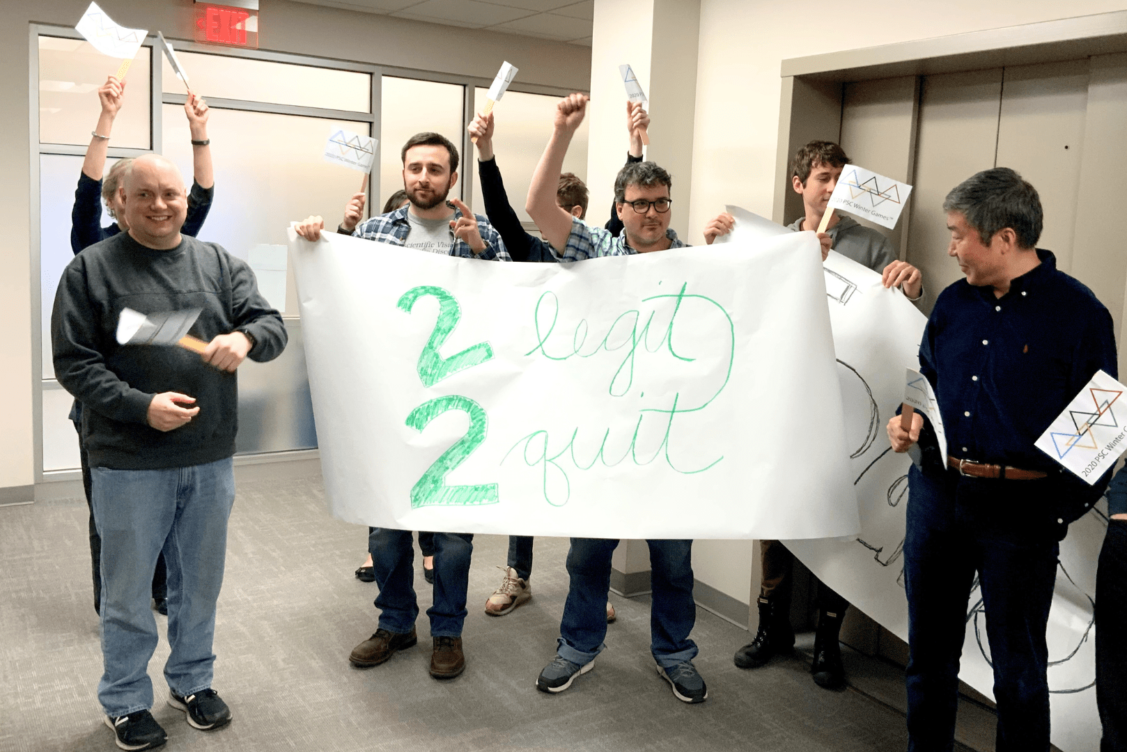 A group of people in an office wave makeshift flags and hold a large banner that reads "2 legit 2 quit"